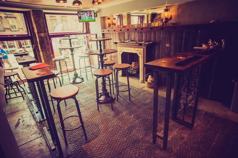 Interior of the Wee Pub, the Smallest pub in Scotland, wiht wooden bar stools around high wooden tables. the floor is stone tiles and the wall is covered with dark wood panelling with a stone fieplace. 