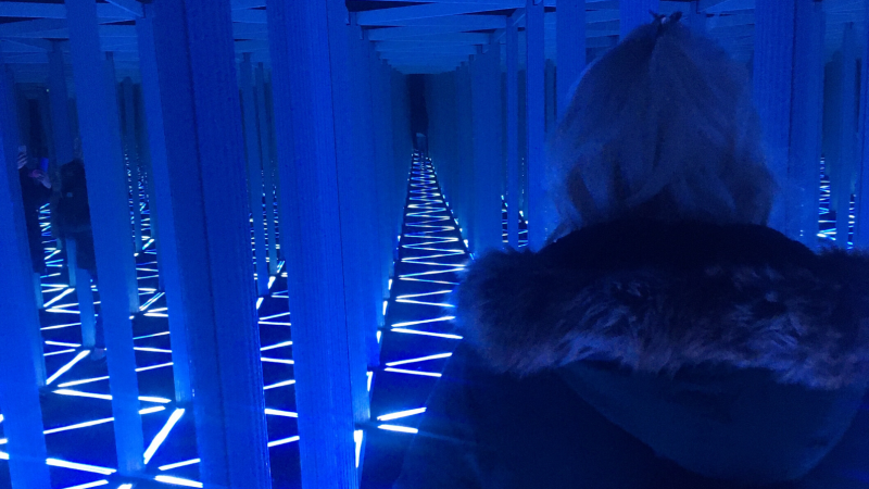 Blonde haired girl wearing a coat with a fur-lined hood inside a narrow mirrored hallway lit with zigzagging blue lights in the Camera Obscura in Edinburgh