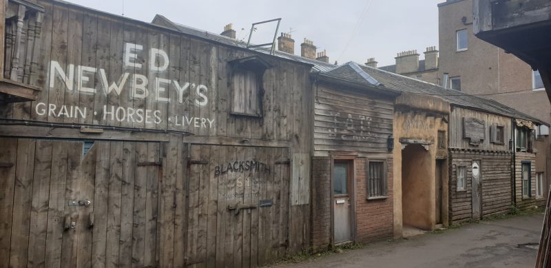 A street in the fake Wild West Ghost Town in edinburgh with small, wooden clapboard style houses. the closes is painted with the words ED Newbey's grain, horses, livery 