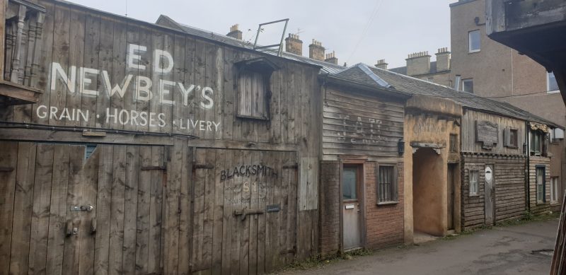 A street in the fake Wild West Ghost Town in edinburgh with small, wooden clapboard style houses. the closes is painted with the words ED Newbey's grain, horses, livery
