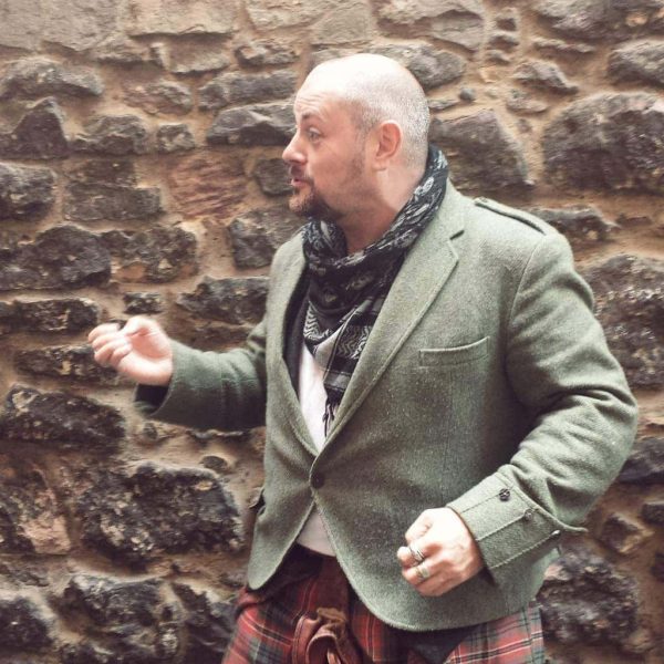 Portrait of a man with a shaved head an light brown goatee wearing a red tartan kilt and a dark greey woolen blazer jacket standing in front of a stone wall and gesturing as he tells a story. Edinburgh off the beaten path