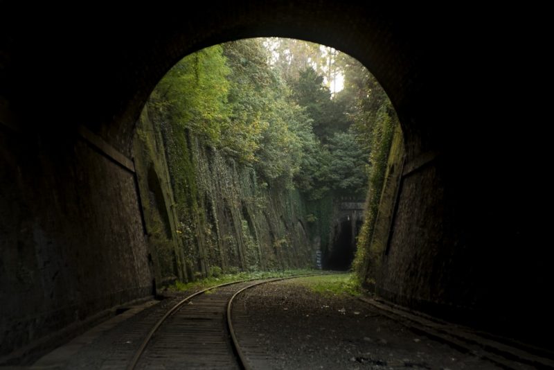 taken from inside an old stone train tunnel looking out where the curved train track is running past a tall stone wall with green leafy trees above