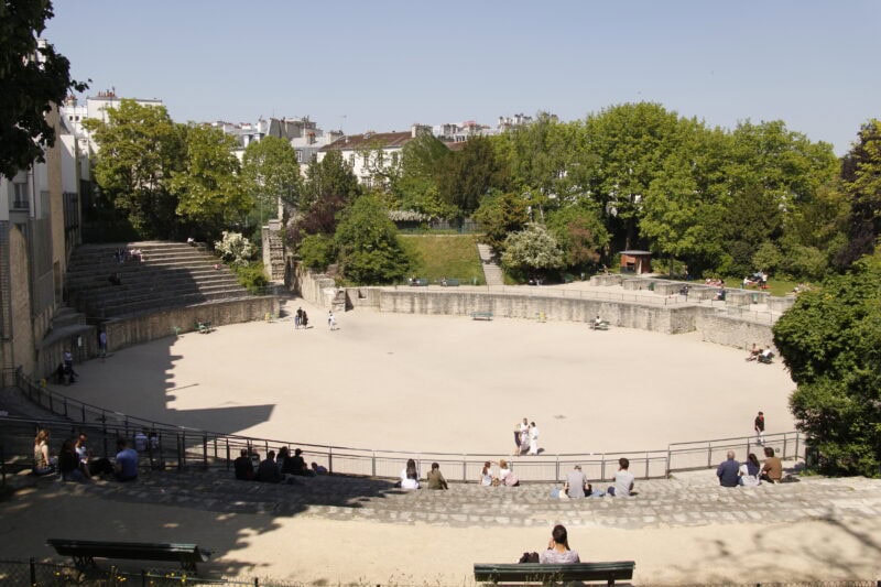 Looking down at the circular space in the centre of Arènes de Lutèce,surrounded by a low grey stone wall with rows of curved ampitheatre style seating on the right side.