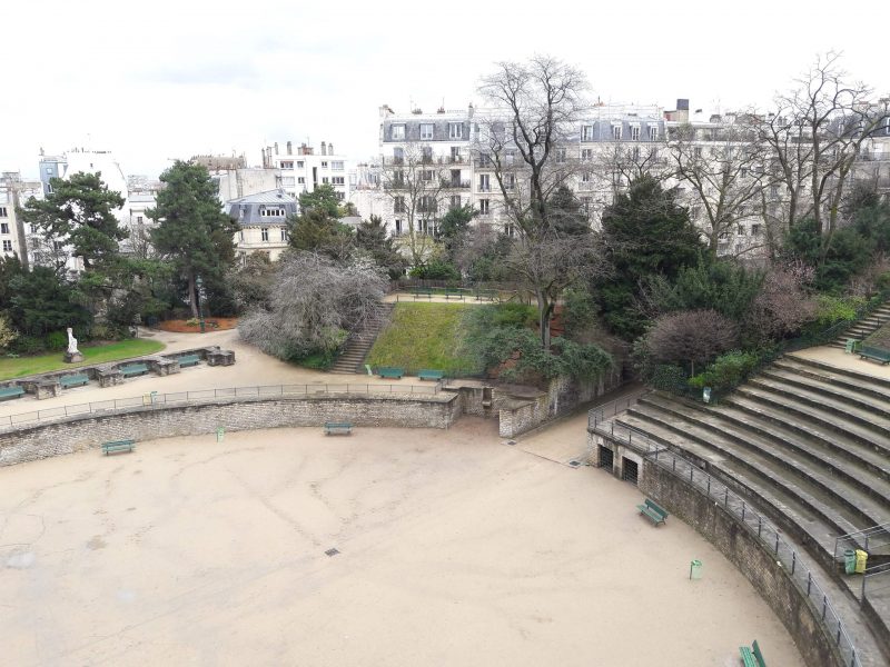 Looking down at the circular space in the centre of Arènes de Lutèce,surrounded by a low grey stone wall with rows of curved ampitheatre style seating on the right side. the arena is surrounded by a park with most of the trees bare in winter except a few leafy evergreens. there are tall white Parisian townhouses in the distance and an overcast sky. Unusual things to do in Paris. 