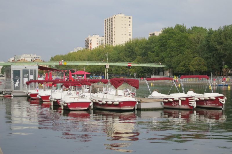 Line of small red and white motorboats on the River Siene with red canapoies, lined up along a floating pontoon with a grey ticket office on it. there is a narrow green brideg crossing the river behind and a thick row of leafy trees on the far bank. Unusual things to do in Paris. 