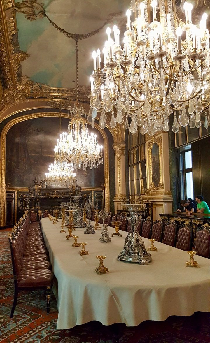 Interior of the Foreign Affairs Ministry in Paris, a large dining room with a very long table beneath a high ciling. the table has a white tablecloth and many small golden chalices with tall silver candlestick holders in the centre and is surrounded by brown leather dining chairs. There are three crystal chandeliers above and the far wall has a large painting with the detail hidden by the light from the chandelier. the edges of the wall and cieling are painted gold. 