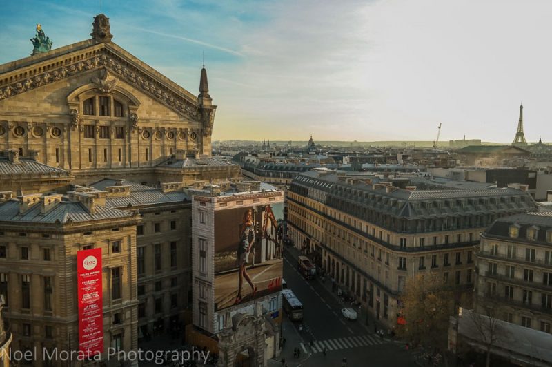 Rooftop view from Galeries Lafayette with a large, historic stone building opposite with a triangular roof that has a caved facade and the rest of the paris city skyline beyond with the eiffel tower in the distance against an almost-white sunset sky