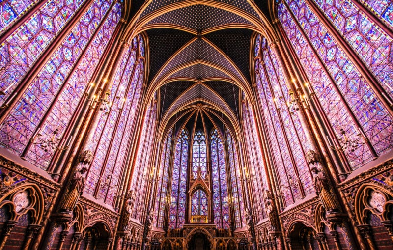 interior of St Chapelle church in Paris with stone vaulted ceiling and every wall covered with floor to cieling stained glass in different shades of pink and purple