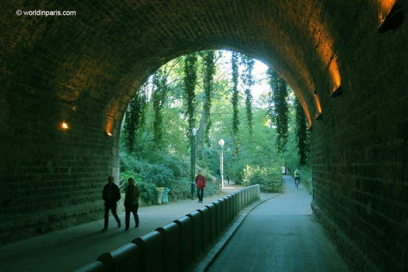 Looking out of an old stone railway tunnel where the tracks have been replaced by paths and a bike lane, there are two people walking into the tunnel silhouetted against the background and a man in a red jacket walking behind them about to enter the tunnel. Outside the tunnel the paths run through a forest of green leafy trees. The Coulee Verte Paris