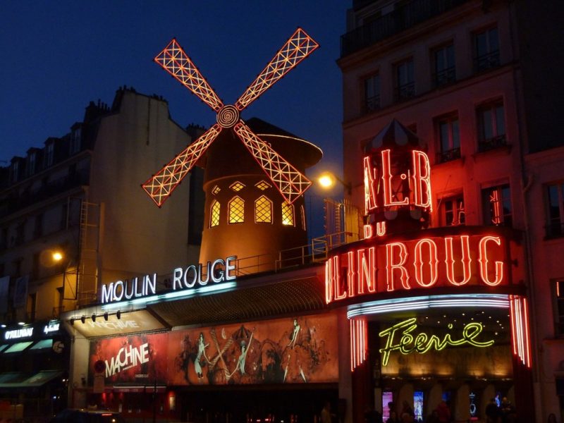 exterior of the Moulin Rouge nightclub in Paris at night with a neon sign with the name below a small windmill lit in yellow wiht its arms covered in red neon lights. To the right of this is the entrance with a large curved sign with red neon letters saying Moulin Rouge