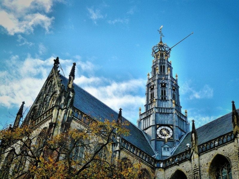 A Gothic-style church with a prominent clock tower, detailed with pointed arches, spires, and a large clock face. Autumn-colored trees are in the foreground under a partly cloudy sky.