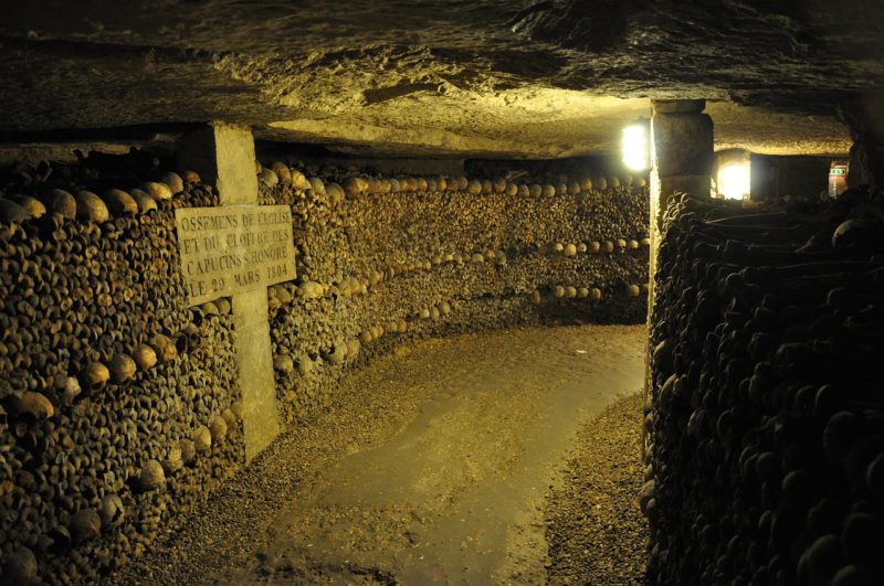 underground tunnel in the Paris Catacombs with a stone path dimly lit by a single yellow electric light, the left side of the tunnel is a curving wall made completely from skulls and bones designed around a large stone cross.