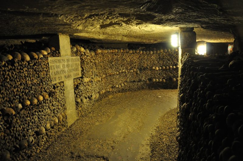 underground tunnel in the Paris Catacombs with a stone path dimly lit by a single yellow electric light, the left side of the tunnel is a curving wall made completely from skulls and bones designed around a large stone cross. 