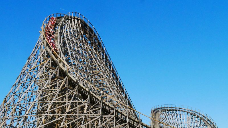 "A large wooden roller coaster at Efteling theme park in the Netherlands with a steep drop, against a clear blue sky. The train full of riders is descending the slope. The structure is made up of numerous wooden beams forming a complex lattice framework.