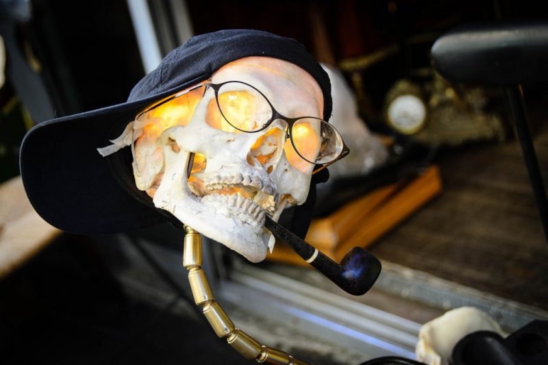 model of a human skull on an old metal lamp with the bulb inside the skull. the skull is wearing glasses and a sideways black baseball cap and has a pipe in its mouth. on a stall at a flea market. 