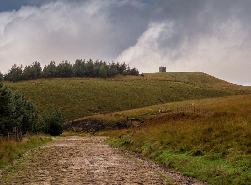 A scenic landscape with a dirt path leading up to a grassy hill known as Rivington Pike, topped with a small stone tower. The sky is cloudy, with a mix of dark and light clouds.