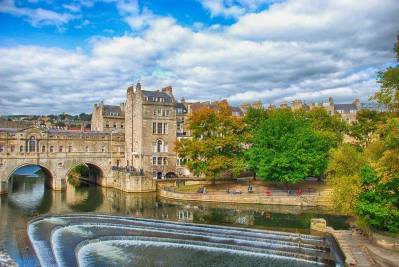 wide river in Bath England with a three-layered semi-circular weir across it in front of a walled stone bridge built from beige stone. Pulteney Bridge in Bath