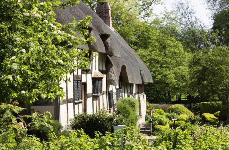 A charming thatched-roof cottage in Stratford Upon Avon, surrounded by a lush, well-kept garden. The timber-framed structure has white plaster walls and small, leaded glass windows. The garden is filled with various shrubs, bushes, and trees.