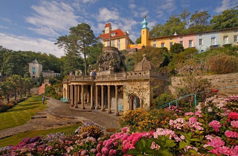 flowery garden with lots of pink flowers and amn italianate collonaded stone structure art the base of a very low cliff with the tops of a few colourful builings of Portmeirion in the background