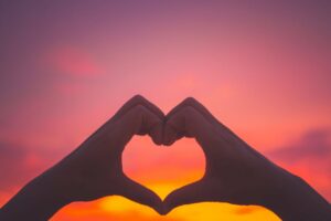 Silhouette of two hands creating a heart shape in front of a pink and orange sunset sky. Romantic Places in the UK.