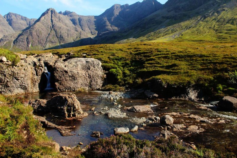 A small waterfall flowing over rocks into a shallow stream with grassy banks and towering mountains in the background, on the Isle of Skye in Scotland