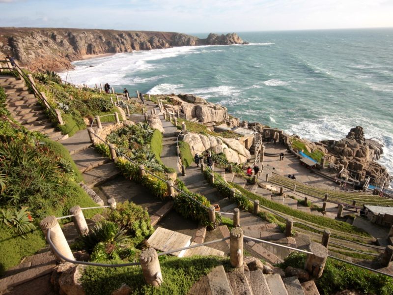 View of the Minack Theatre in Cornwall, an open air theatre built from beige stone on a cliff edge with the coastline stretching away beyond.