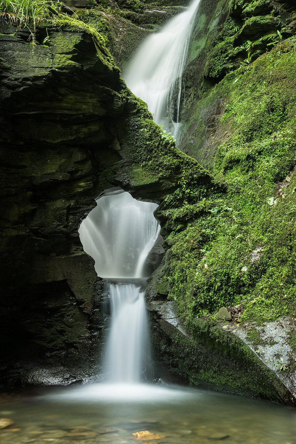 A serene waterfall flowing through a moss-covered rock formation with a natural archway, surrounded by lush greenery - St Nectan's Glen Cornwall.