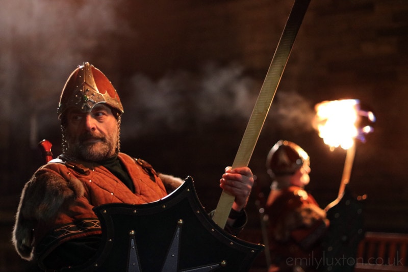 Man dressed as a viking with red tunic and a red and gold helmet holding a wooden torch and and a sheid at night in the Edinburgh Hogmanay Torchlight Procession