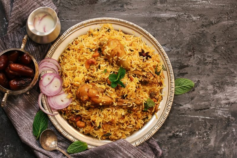 flat lay of a bowl of yellow rice with chicken legs and red onion next to a silver bowl of dates and a silver jug of white sauce.