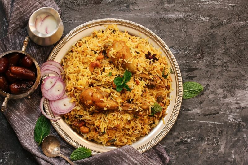 flat lay of a bowl of yellow rice with chicken legs and red onion next to a silver bowl of dates and a silver jug of white sauce. 