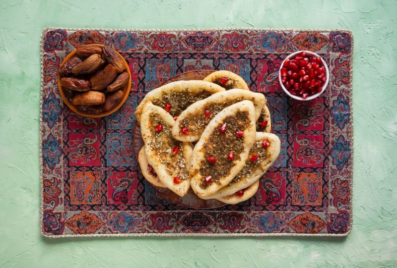 flatlay of a colourful middle eastern style rug with a plate of flatbreads on it, each of them topped with herbs and pomegranite seeds, next to a bowl of dates and a bowl of pomegranite seeds. dubai food guide
