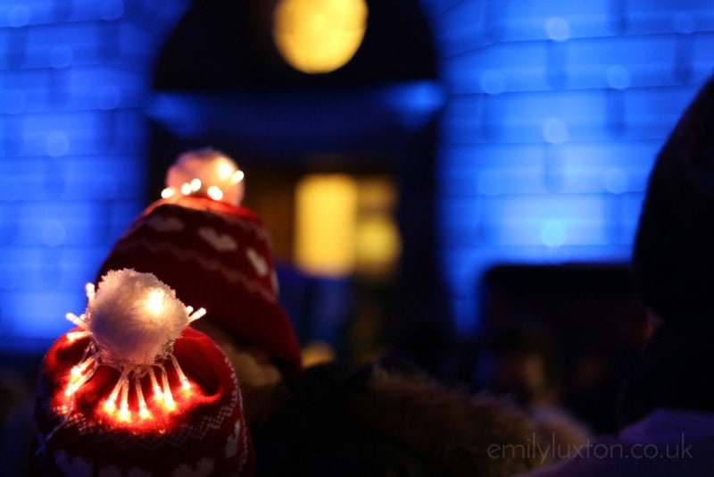 back of a head wearing a red and white wool hat with a white pompom covered in fairy lights standing in a dark street at night with a blue-lit stone wall behind