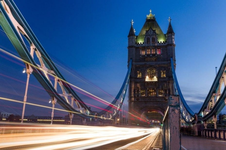 shot of tower bridge in london at night with a long exposure making the car lights blur as they cross the bridge. the tower is mostly dark with a few lights on its facade against a clear, dark blue night sky. London is one of the best cities for nightlife in the uk.