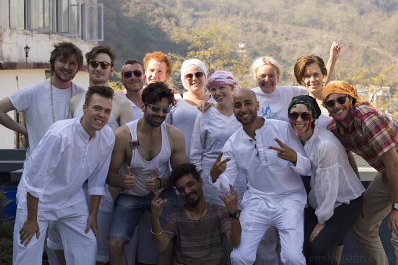 Group of men and women huddled together on a roof terrace for a photo, there is a view of a tree covered hill behing, and everyone is wearing white. Holi India survival tips