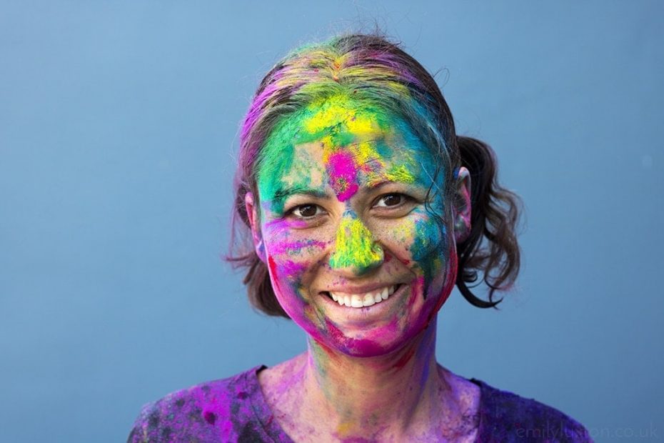Close up of a woman's face, she has white skin and short brown hair and is smiling at the camera, and her face and hair are covered with different coloured paint. Girl's Guide to Holi