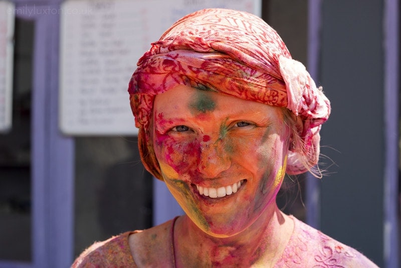 Close up of a white woman's face covered in different coloured paint, she is wearing a hewadscarf and kaftan that have been stained pink and orange by the dye.