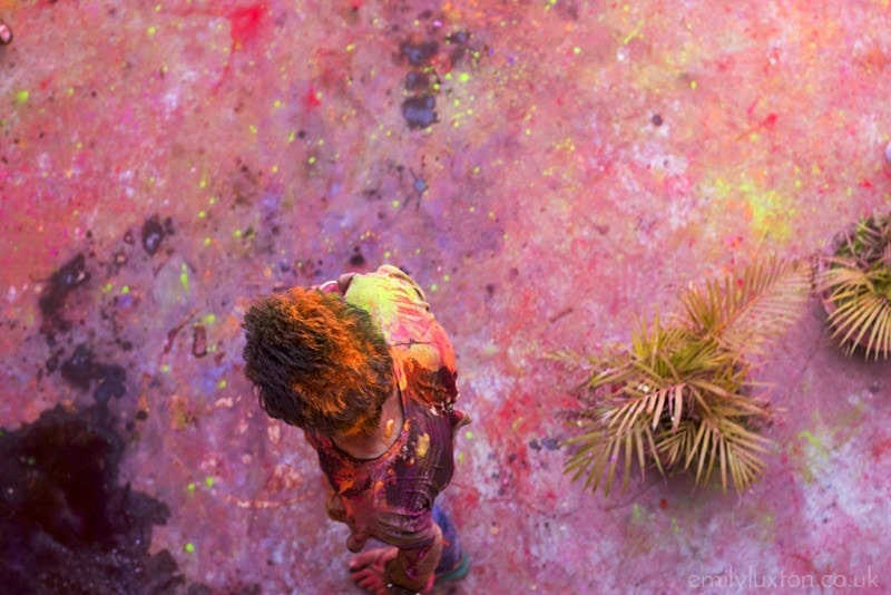 Aerial shot looking down at an indian man covered in paint standing in a courtyard where the concrete floor is splattered with different coloured paint.