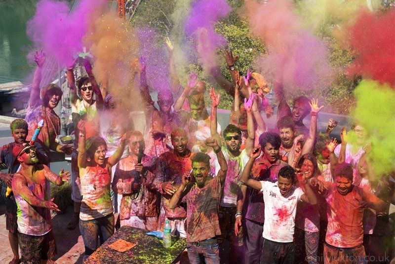 Group of men and women standing close together and throwing different coloured powders into the air at Holi. 