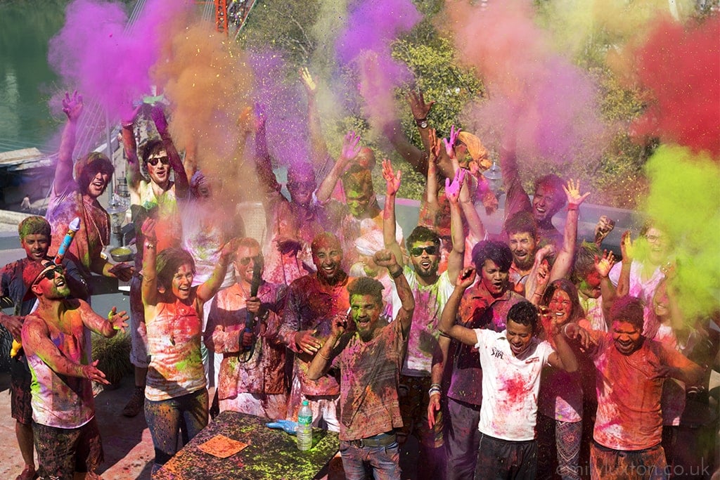 Group of men and women standing close together and throwing different coloured powders into the air at Holi.