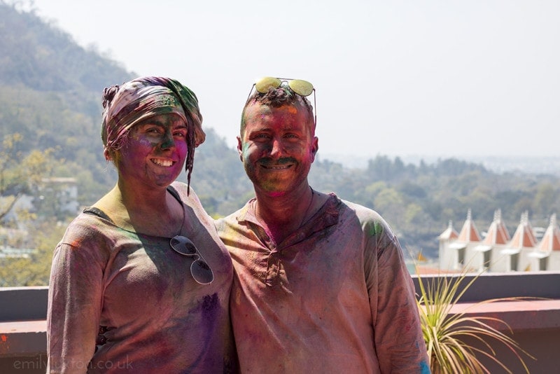 Emily standing next to a man on a roof terrace with a wooded hillside behind, both are wearing loose long sleeved tops stained purple and red by paint and both have multicoloured paint all over their faces. Emily is also wearing a headscarf stained in many different colours. 