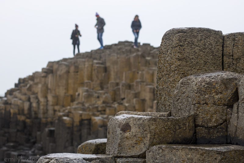 Close up of several hexagonal basalt columns on the Giant's Causeway in Northern Ireland UK wiht more rock columns out of focus behind and three figures walking across the top of them