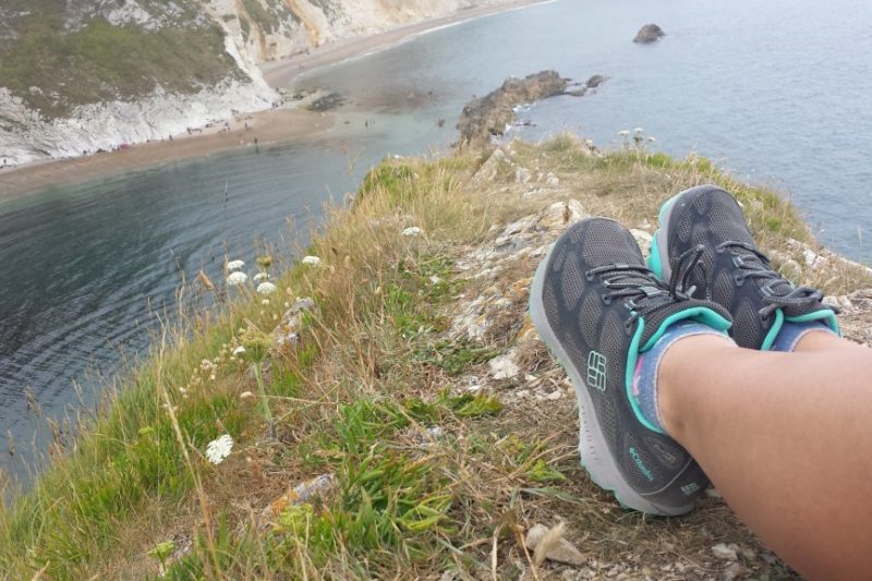Feet in grey trainers on a grassy cliff edge with the sea below and more cliffs in the distance