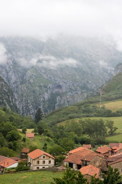 mountain valley with a small village of red roofed houses amongst green hills with a rocky mountain behind and misty clouds above. best national parks in spain. 