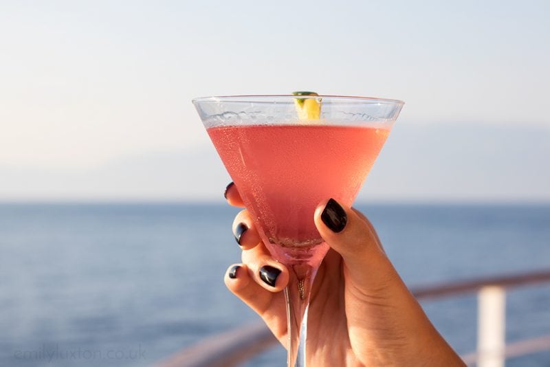 hand holding a pink cosmo cocktail in a conical cocktail glass with the ocean behind on the edge of a cruise ship. tips for taking a solo cruise