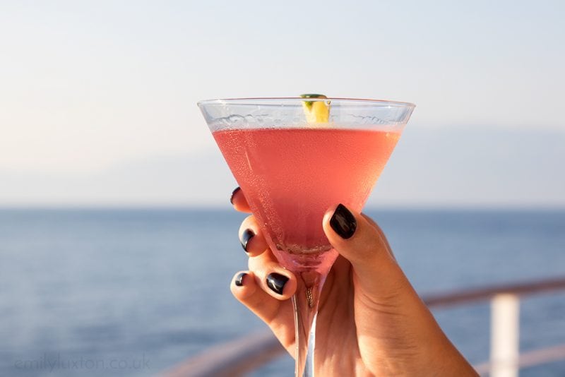 hand with black painted nails holding a pink cosmo cocktail in a small cone shaped glass with a wedge of lime in front of the blue sea