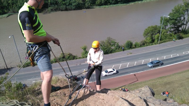 a girl in black leggings, white fleece jumper and yellow helmet is abseiling over a cliff above a road beside a river at Avon Gorge Bristol - UK adventure 