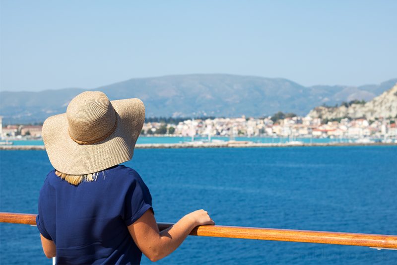 emily wearing a navy blue t shirt and grey floppy straw hat standing at a railing on a cruise ship looking away from the camera towards the blue sea and distant white cliffs in greece. tips for taking a solo cruise