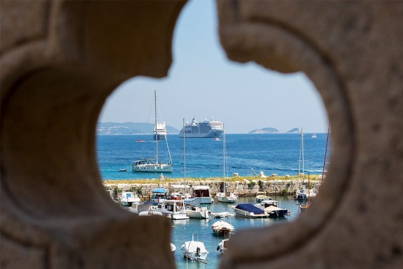 cross shaped window in a stone wall with a view of a small marina filled with sailing boats along a stone harbour wall, with the blue sea beyond and a large cruise ship moored there.