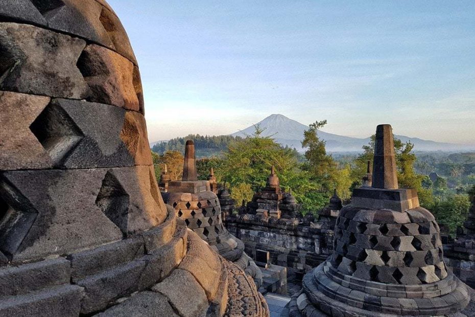 stone domes at Borobadur in Yogyakarta just after sunrise with a view of a conical mountain in the distance and blue sky overhead