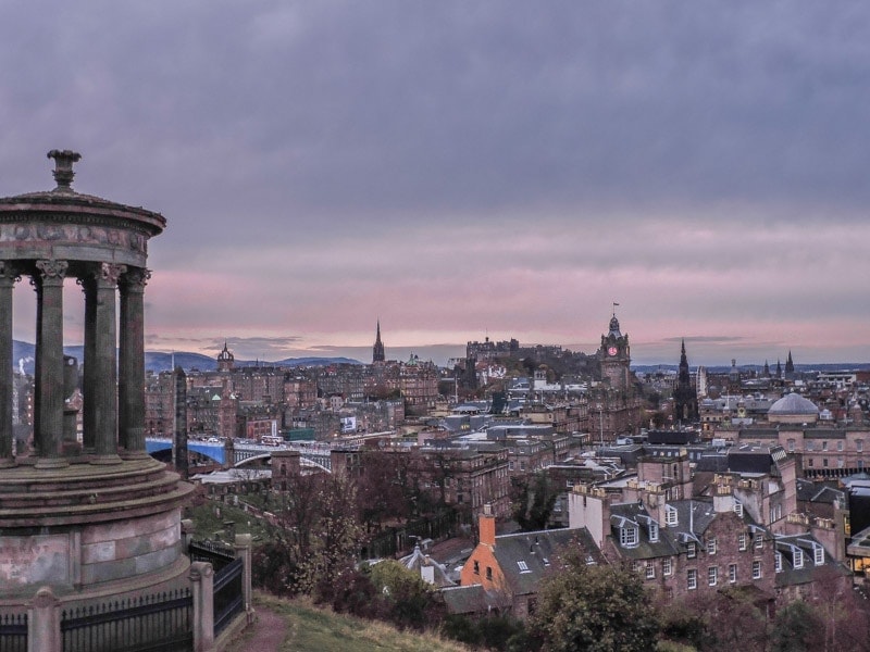 circular stone monument with columns on a hilltop with a view of edinburgh city skyline behind at dusk with a pink and purple sky in winter